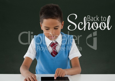 Happy student boy at table using a tablet against green blackboard with back to school text