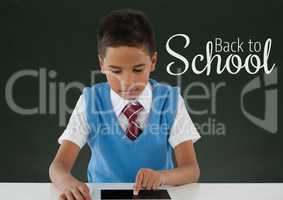 Happy student boy at table using a tablet against green blackboard with back to school text