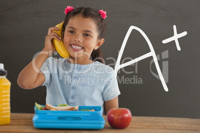 Happy student girl at table holding a banana against grey blackboard with A+ text