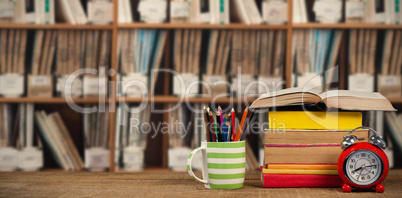 Composite image of stack of books by mug with colored pencils and alarm clock on wooden table