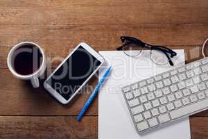 Mobile phone, keyboard, paper, spectacles, pen and coffee on wooden table