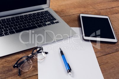 Digital tablet, laptop, paper, pen and spectacles on wooden table