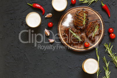 Tasty barbecue steaks and beer in glasses on a black stone background, with cherry tomatoes and chili peppers