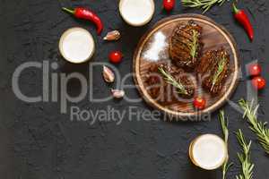 Tasty barbecue steaks and beer in glasses on a black stone background, with cherry tomatoes and chili peppers