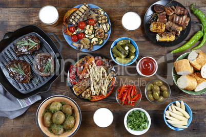 Dinner table with meat grill, bbq vegetables, salads, sauces, snacks and beer, top view