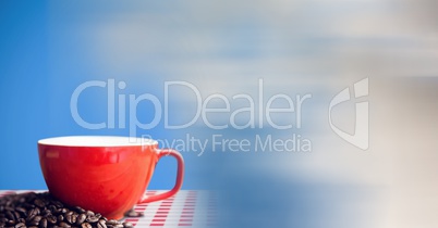 Red coffee cup on red and white table cloth with beans against blue background and blurry sky transi