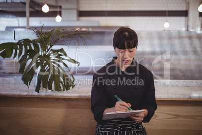 Young waitress writing on clipboard while sitting against counter