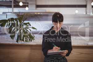 Young waitress writing on clipboard while sitting against counter