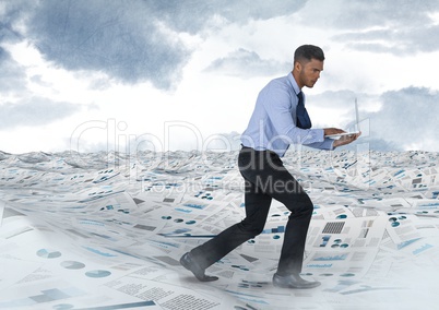 Businessman on laptop in sea of documents under sky clouds