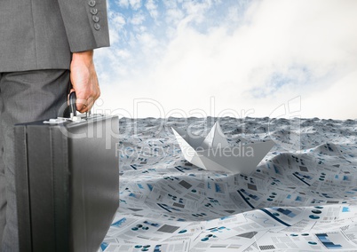 Businessman holding briefcase in sea of documents with paper boat