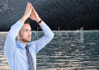 Business man meditating against water and trees on hill