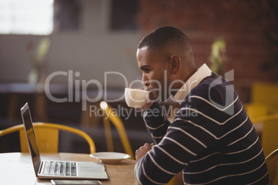 Side view of young man drinking coffee while looking at laptop