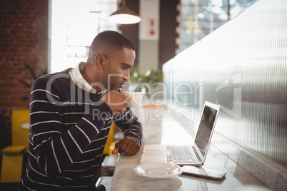 Side view of young man drinking coffee while looking at laptop