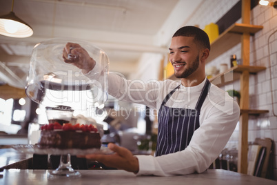 Smiling young waiter holding glass lid over cakestand at counter