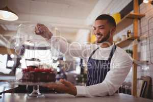 Smiling young waiter holding glass lid over cakestand at counter