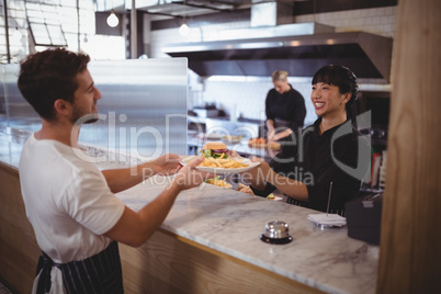Smiling young female chef giving burger and fries to waiter