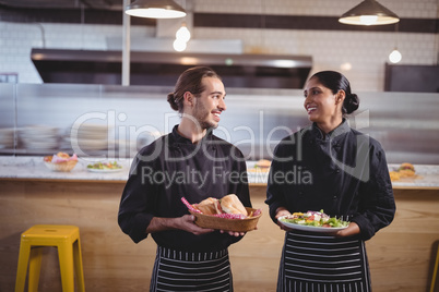 Smiling young wait staff looking at each other while holding fresh food in coffee shop