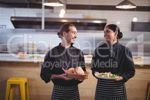 Smiling young wait staff looking at each other while holding fresh food in coffee shop