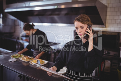 Young waitress talking on smartphone while waiter preparing food in commercial kitchen