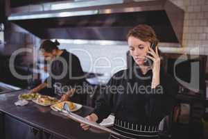 Young waitress talking on smartphone while waiter preparing food in commercial kitchen