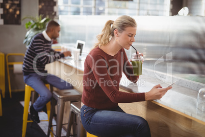 Young blond woman using smartphone while drinking juice at counter