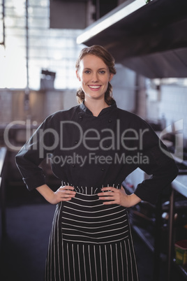 Portrait of smiling young female barista standing with hands on hip in kitchen