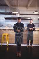 Portrait of confident young wait staff in uniform standing with arms crossed against counter