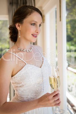 Bride holding champagne looking through window at home