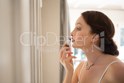 Beautiful bride applying lipstick while looking into mirror at home