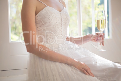 Midsection of bride holding champagne while sitting by window