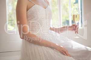 Midsection of bride holding champagne while sitting by window