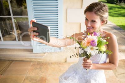 Happy bride holding bouquet taking selfie with mobile phone while sitting on chair in yard