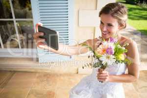 Happy bride holding bouquet taking selfie with mobile phone while sitting on chair in yard