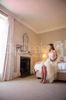 Thoughtful bride holding champagne looking away while sitting on bed at home
