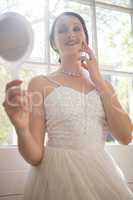 Attractive bride looking into hand mirror while standing by window at home