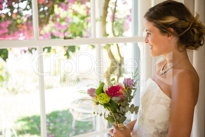 Side view of smiling beautiful bride holding bouquet while looking through window