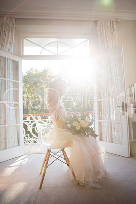 Beautiful bride holding bouquet while sitting on chair at home