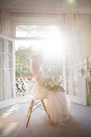 Beautiful bride holding bouquet while sitting on chair at home