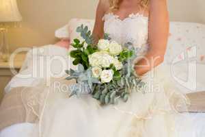 Midsection of bride in wedding dress holding bouquet while sitting on bed