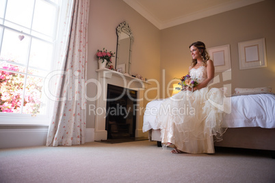 Beautiful bride holding bouquet while sitting on bed at home