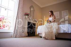 Beautiful bride holding bouquet while sitting on bed at home