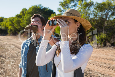 Young woman looking through binoculars while boyfriend drinking water in forest