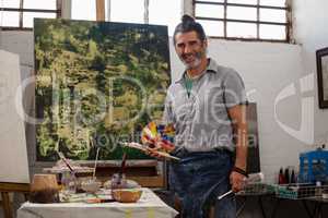 Portrait of confident man standing with palette and paintbrushes