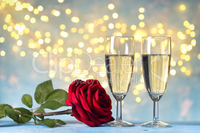 Two champagne glasses and red rose flower, against holiday lights