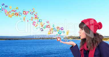 young woman with hat blowing application icons from her hands in front of the sea and mountains
