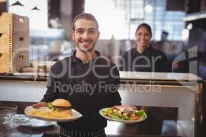 Portrait of smiling young waiter serving food while standing against waitress