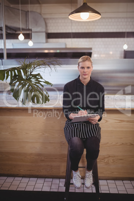 Portait of confident young waitress sitting with clipboard at coffee shop