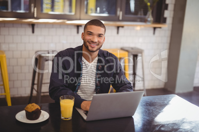 Portrait of smiling young man sitting with laptop at coffee shop