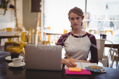 Confident woman writing in dairy while sitting with laptop at coffee shop