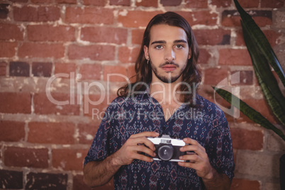 Portrait of confident young photographer holding camera against brick wall